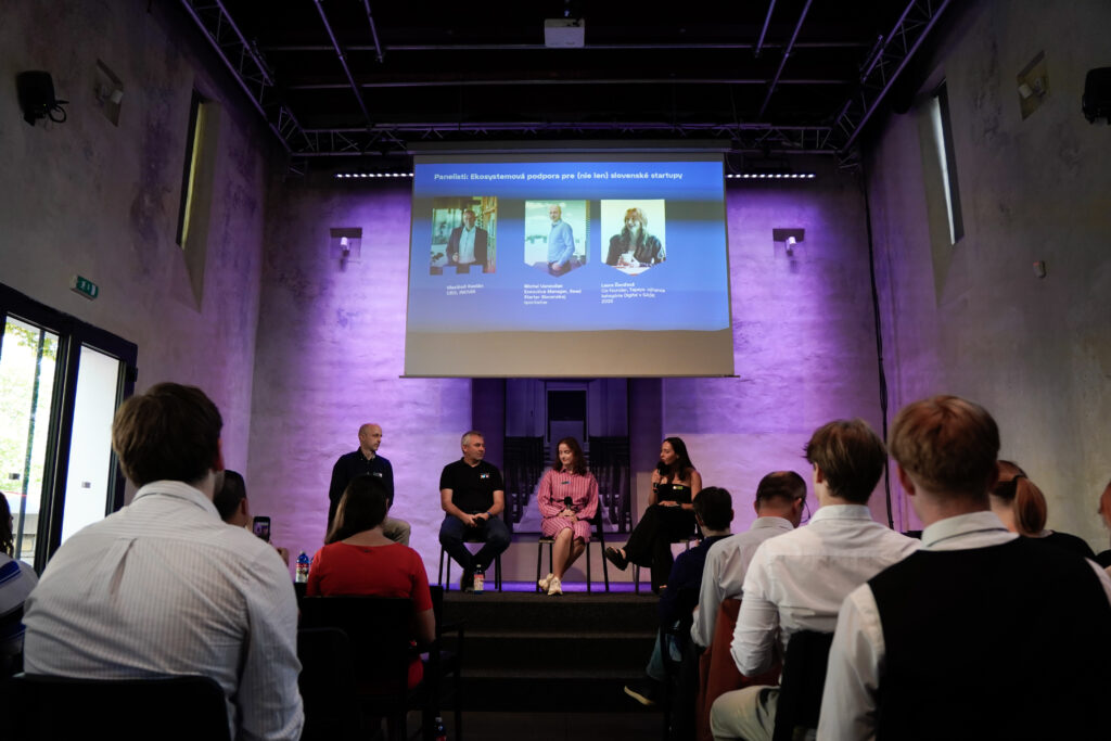 Publikum sediace chrbtom sleduje panelovú diskusiu na pódiu fialovo nasvietenej Novej Synagógy v Žiline. Na pódiu štyria ľudia, nad nimi plátno s fotkami panelistov.